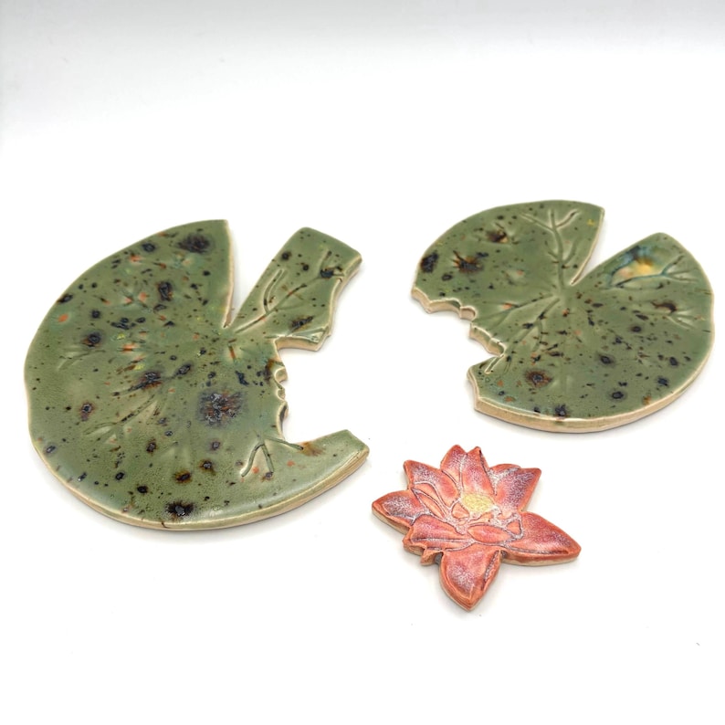 May include: Ceramic lily pad and lotus flower set. The lily pads are green with brown speckles and leaf vein details. The lotus flower is pink with yellow and orange accents.