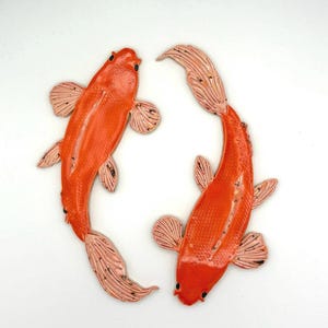 May include: Two ceramic koi fish sculptures in a vibrant orange hue, with delicate pink and black accents on their fins and tails. The fish are arranged in a circular pattern, showcasing their detailed craftsmanship.
