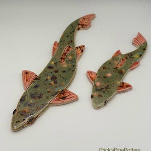 May include: Two ceramic fish figurines. The fish have a speckled olive green body with pink fins and tail. The fish are different sizes and are positioned on a white background. The artist's name, PricklyPinePottery, is in the bottom right corner.