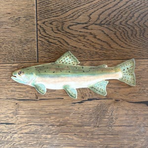 May include: A decorative fish figurine with a detailed painted design. The fish features a light green body with darker green spots, a white underbelly, and blue-green fins. The figurine is lying on a wooden surface.