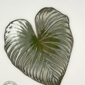 May include: A ceramic leaf-shaped decorative piece with a glossy, dark green finish. The leaf features detailed veining and a slightly raised edge. The Prickly Pine Pottery logo is visible in the lower left corner. The piece is set against a white background.