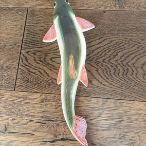May include: A wooden fish figurine with a green and brown back, white belly, and pink fins and tail. The fish is lying on a wooden surface.
