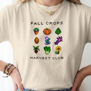 May include: A beige t-shirt featuring a pixel art graphic of various autumn vegetables and flowers arranged in a grid. The text "FALL CROPS" appears above the grid, and "HARVEST CLUB" is printed below in a black, blocky, pixelated font.