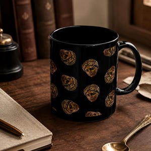 May include: A black ceramic mug featuring a repeating pattern of small, golden-toned classical sculpture faces. The mug has a smooth, glossy finish, a cylindrical body, and a curved handle on the right side. The golden faces are depicted in relief.