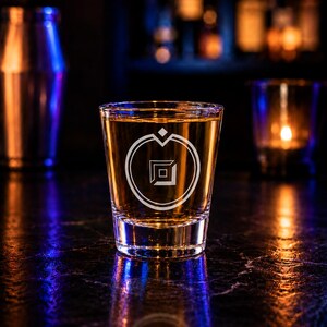 May include: A clear glass shot glass filled with an amber-colored liquid. The front of the glass features a white etched circular logo with a diamond shape and a small square in the center. The glass has a thick base and straight sides.