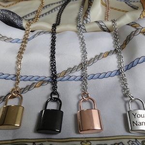 May include: Four padlock pendant necklaces in gold, black, rose gold, and silver. The silver padlock has the text "Your Name" engraved on it. Each necklace has a matching chain and is displayed on a white fabric with a nautical rope design.