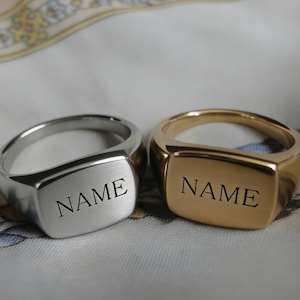 May include: Two signet rings, one silver and one gold, each with the word "NAME" engraved on the rectangular face. The rings are displayed on a patterned fabric, showcasing the contrast between the metals.