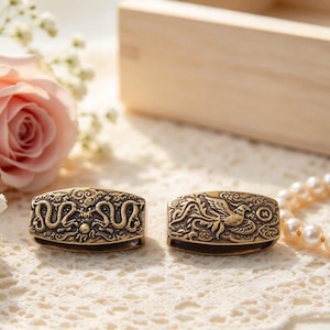 May include: Two rectangular brass beads featuring intricate embossed designs. One bead displays a detailed dragon motif with swirling clouds, while the other bead showcases a phoenix figure surrounded by ornate patterns and a small circular emblem.