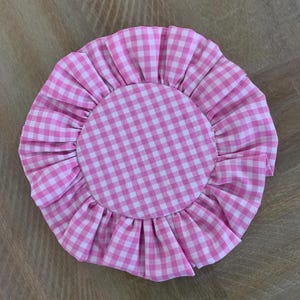 Op de afbeelding: Een rond stoffen item met een roze en witte gingham patroon, met een gerimpelde rand. Het item heeft een centraal gingham ontwerp en een gerimpelde rand. De stof toont een traditioneel roze en wit ruitpatroon.