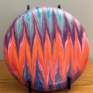 May include: A round, colorful decorative piece with a vibrant abstract design. The artwork features a blend of orange, pink, purple, and blue hues, creating a dynamic and eye-catching visual effect. The piece is displayed on a black metal stand.