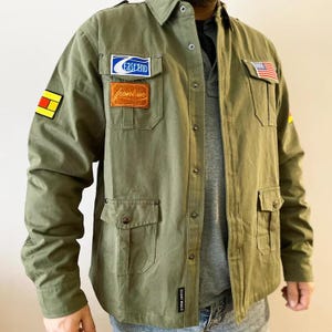 May include: An olive green jacket with multiple pockets, patches, and a button-up front. Features a small American flag patch and the text "Fastend" and "Frontier" on the chest. The jacket has a collared neckline and long sleeves.