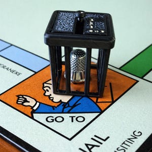 Pu&ograve; includere: Un pezzo del gioco del Monopoli raffigurante una cella di prigione nera con un ditale d'argento all'interno, posizionato sulla casella "Vai in prigione" di un tabellone del Monopoli. Il tabellone ha sezioni arancioni, blu e verdi.