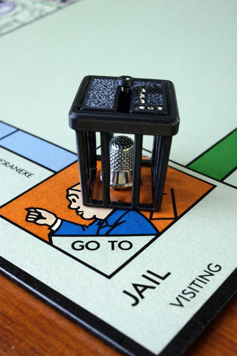 Pu&ograve; includere: Un pezzo del gioco del Monopoli raffigurante una cella di prigione nera con un ditale d'argento all'interno, posizionato sulla casella "Vai in prigione" di un tabellone del Monopoli. Il tabellone ha sezioni arancioni, blu e verdi.