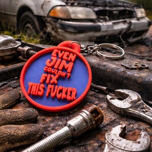 May include: A red and blue keychain with the text "EVEN JIM COULDN'T FIX THIS FUCKER" sits among tools and a damaged car with the hood open. The keychain is round with a silver chain.