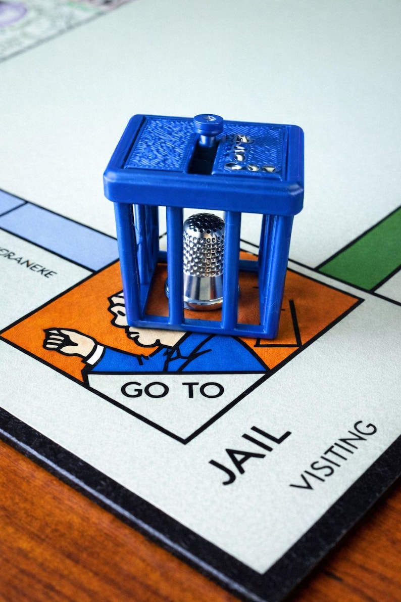 Pu&ograve; includere: Un pezzo del gioco in plastica blu a forma di cella di prigione con un ditale d'argento all'interno, posto su un tabellone di gioco del Monopoli. Il tabellone mostra la casella "Vai in prigione".