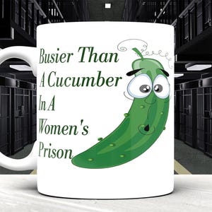 Könnte beinhalten: Weiße Keramiktasse mit dem Text "Busier Than A Cucumber In A Women's Prison" und einer Cartoon-Gurken-Illustration. Die Tasse hat einen weißen Henkel und ist vor dem Hintergrund von Gefängniszellen platziert.