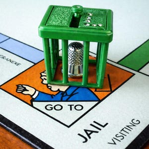 Pu&ograve; includere: Una scena del gioco da tavolo Monopoly con una prigione di plastica verde e un segnaposto a forma di ditale argentato. La casella del tabellone recita "Vai in prigione". Il tabellone di gioco &egrave; visibile sullo sfondo.