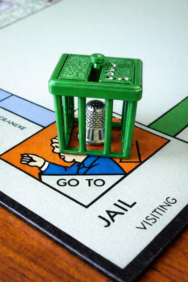 Pu&ograve; includere: Una scena del gioco da tavolo Monopoly con una prigione di plastica verde e un segnaposto a forma di ditale argentato. La casella del tabellone recita "Vai in prigione". Il tabellone di gioco &egrave; visibile sullo sfondo.