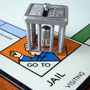 Pu&ograve; includere: Un segnaposto del gioco del Monopoli color argento raffigurante una cella di prigione con un ditale all'interno, posizionato sulla casella "Vai in prigione" di un tabellone del Monopoli. Il tabellone &egrave; verde chiaro, con sezioni arancioni, blu e verdi.