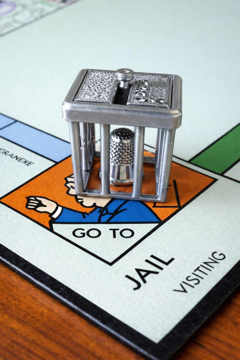 Pu&ograve; includere: Un segnaposto del gioco del Monopoli color argento raffigurante una cella di prigione con un ditale all'interno, posizionato sulla casella "Vai in prigione" di un tabellone del Monopoli. Il tabellone &egrave; verde chiaro, con sezioni arancioni, blu e verdi.