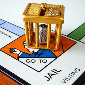 Pu&ograve; includere: Una gabbia in metallo color oro con un ditale d'argento all'interno, posta su un tabellone di gioco del Monopoli. Il tabellone mostra lo spazio "Vai in prigione", con una figura in abito blu. La gabbia ha una parte superiore decorativa.