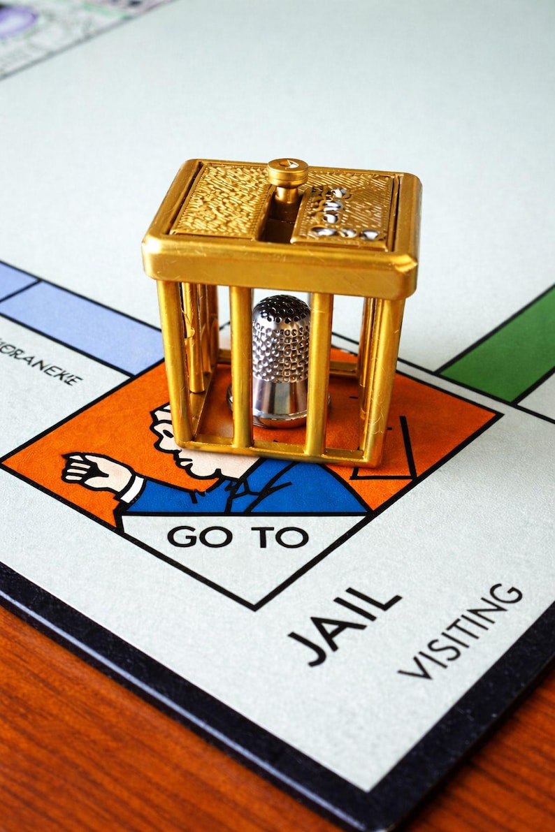 Pu&ograve; includere: Una gabbia in metallo color oro con un ditale d'argento all'interno, posta su un tabellone di gioco del Monopoli. Il tabellone mostra lo spazio "Vai in prigione", con una figura in abito blu. La gabbia ha una parte superiore decorativa.