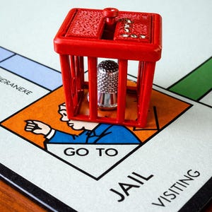 Pu&ograve; includere: Una cella di prigione di plastica rossa con un ditale d'argento all'interno, appoggiata su un tabellone di gioco del Monopoli. Il tabellone mostra lo spazio "Vai in prigione". Il pezzo del gioco &egrave; un elemento classico del gioco.