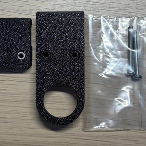 Op de afbeelding: Een set zwarte, getextureerde metalen hardware componenten, waaronder een klein vierkant stuk met twee gaten, een groter stuk met een ring en een doorzichtige plastic zak met twee schroeven. De hardware is waarschijnlijk voor montage of assemblage.