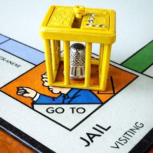 Pu&ograve; includere: Un pezzo del gioco in plastica gialla a forma di cella di prigione con un ditale d'argento all'interno, posto su una tavola di Monopoly. Il tabellone mostra lo spazio "Vai in prigione".