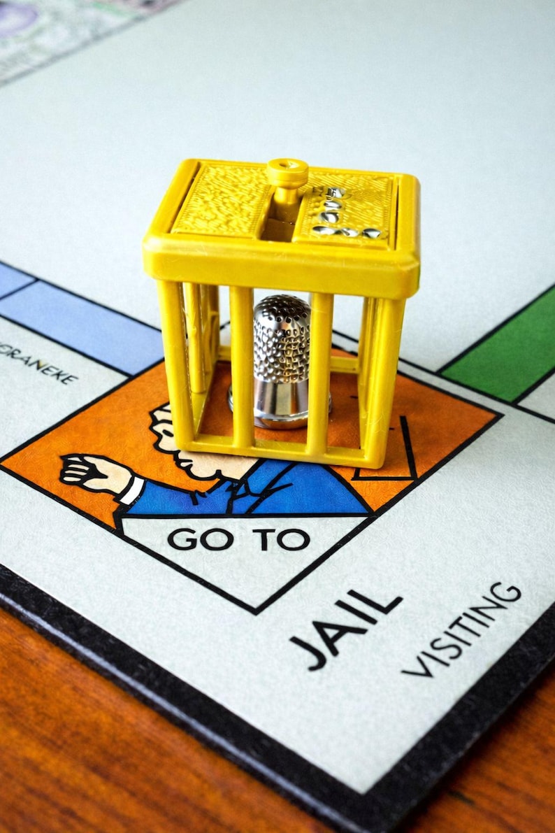 Pu&ograve; includere: Un pezzo del gioco in plastica gialla a forma di cella di prigione con un ditale d'argento all'interno, posto su una tavola di Monopoly. Il tabellone mostra lo spazio "Vai in prigione".