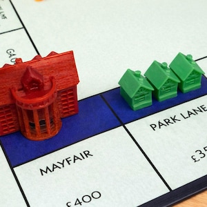 May include: A Monopoly board game scene featuring a red hotel and three green houses on the Park Lane property. The Mayfair property is also visible. The game board is white with black borders and blue property spaces.