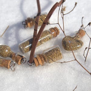 May include: A collection of small glass bottles filled with gold glitter, each sealed with a cork stopper. Some bottles have silver loops attached. The bottles are arranged on a white, snowy surface, with a few twigs scattered around.