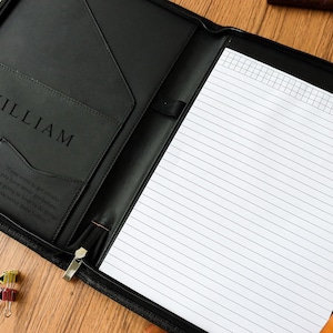 Könnte beinhalten: Schwarzes Leder-Portfolio mit dem eingeprägten Namen "WILLIAM" auf der Vorderseite. Das Portfolio ist geöffnet und zeigt einen linierten Notizblock und das Wort "MEMO" oben. Eine kleine Tasche ist mit den Initialen "WS" und einem Zitat versehen.