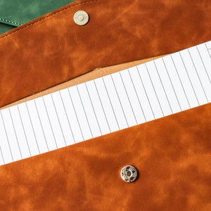 May include: Close-up of a brown leather portfolio with a snap closure, revealing a lined notepad. The portfolio has a textured surface and a contrasting green leather flap is visible in the background. The notepad has a grid pattern.