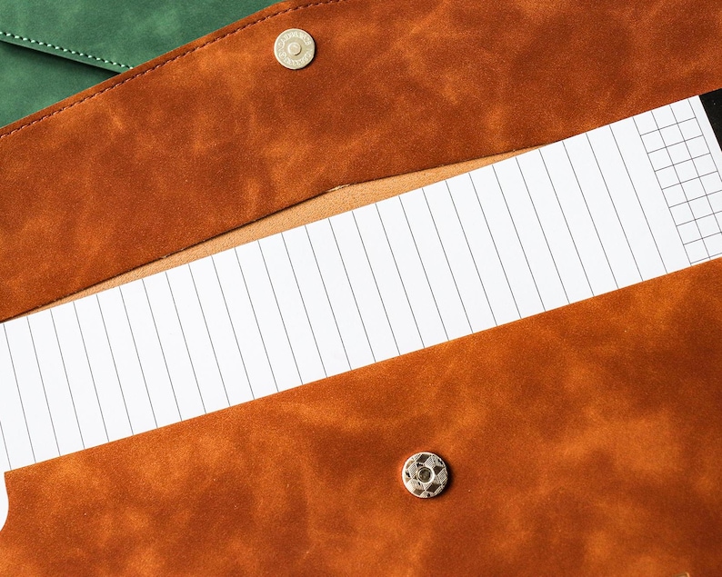 May include: Close-up of a brown leather portfolio with a snap closure, revealing a lined notepad. The portfolio has a textured surface and a contrasting green leather flap is visible in the background. The notepad has a grid pattern.