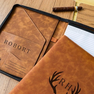 Puede incluir: Un portafolio de cuero marrón con el nombre "ROBORT" grabado en la parte delantera. El portafolio tiene una cremallera negra, un bolsillo y un bloc de notas. También son visibles las iniciales "RH" y un diseño de cornamenta.