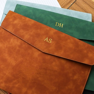 May include: Three personalized leather document holders in shades of brown, green, and blue. Each holder features a flap closure and gold-colored initials. The brown holder is in the foreground, with the initials "AS". The green holder has "DH".