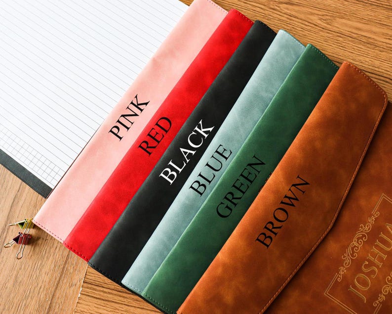 May include: A collection of leather-like notebook covers in pink, red, black, blue, green, and brown. Each cover has a different color and the color name printed on it. A lined notepad is visible on the left.