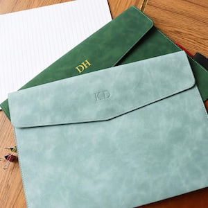 Puede incluir: Portadocumentos de cuero en varios colores, incluyendo verde azulado, verde y marrón. Cada uno tiene un cierre de solapa e iniciales personalizadas. Un bloc de notas con la palabra "MEMO" es visible en el fondo.