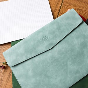 May include: A light teal leatherette document holder with a flap closure, embossed with the initials "KD". A notepad with the word "MEMO" is visible in the background. The document holder is surrounded by other colored document holders.
