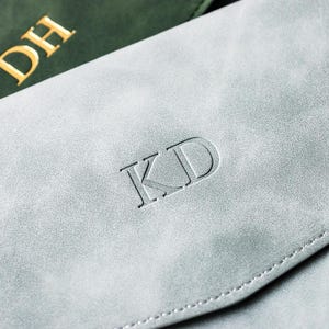 May include: Close-up of two personalized leather items. One is a light gray color with the initials "KD" embossed on the front. The other is a dark green color with the initials "DH" in gold lettering. Both items appear to be a wallet or a similar accessory.
