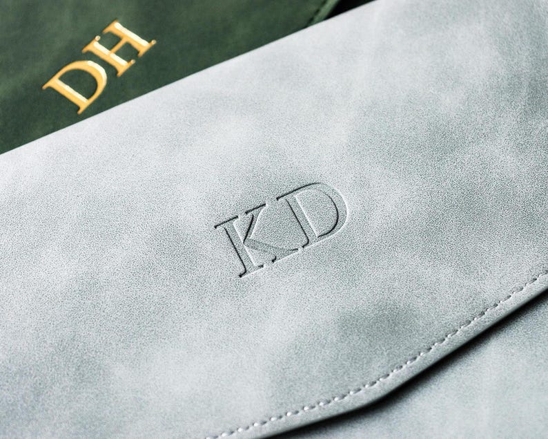 May include: Close-up of two personalized leather items. One is a light gray color with the initials "KD" embossed on the front. The other is a dark green color with the initials "DH" in gold lettering. Both items appear to be a wallet or a similar accessory.