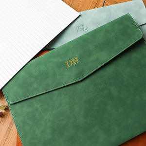 Puede incluir: Portadocumentos de cuero verde, gris y marrón con iniciales personalizadas. El soporte verde tiene "DH" en letras doradas. También se ven un bloc de notas rayado y clips. Los artículos están colocados sobre una superficie de madera.