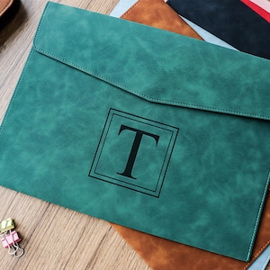 Puede incluir: Una carpeta de documentos de color verde azulado con cierre de solapa, con una letra "T" negra dentro de un marco cuadrado. La carpeta está colocada encima de otras carpetas de cuero de varios colores. Se ve un pequeño clip.