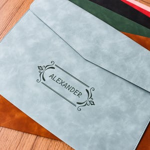May include: A light blue leather portfolio with the name "ALEXANDER" in a decorative frame. The portfolio is on top of a brown leather portfolio, with other colored portfolios visible in the background.