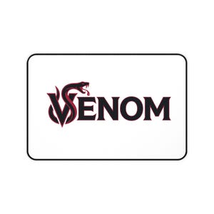 May include: A rectangular white desk mat with a black border. The center features a graphic logo with the word VENOM in bold black letters. The letter V is stylized as a dark red and black snake with its mouth open, integrated into the text.