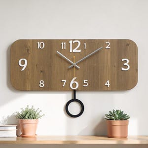 May include: A rectangular wooden wall clock with white numerals and hands. The clock has a pendulum with a black circular weight. The clock is mounted on a light-colored wall, with small potted plants on a shelf below.