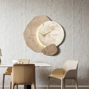 Puede incluir: Un reloj de pared moderno con un diseño único. El reloj presenta un marco beige de forma orgánica con una esfera circular y manecillas doradas. El reloj está montado en una pared blanca texturizada, creando una pieza de decoración elegante.