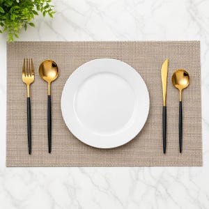 May include: A table setting featuring a white plate, gold-coloured spoon and fork, and a gold-coloured knife. The cutlery has black handles. The setting is on a tan placemat with a grid pattern. A small green plant is in the upper left corner.