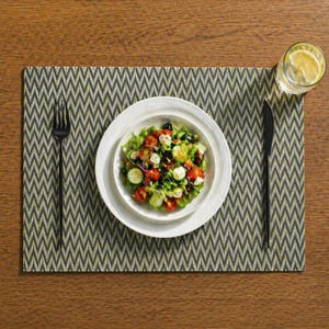 Puede incluir: Una ensalada fresca con tomates, pepinos, aceitunas y queso en un plato blanco, colocada sobre un mantel individual estampado con un tenedor y un cuchillo negros. También hay un vaso de agua con una rodaja de limón.